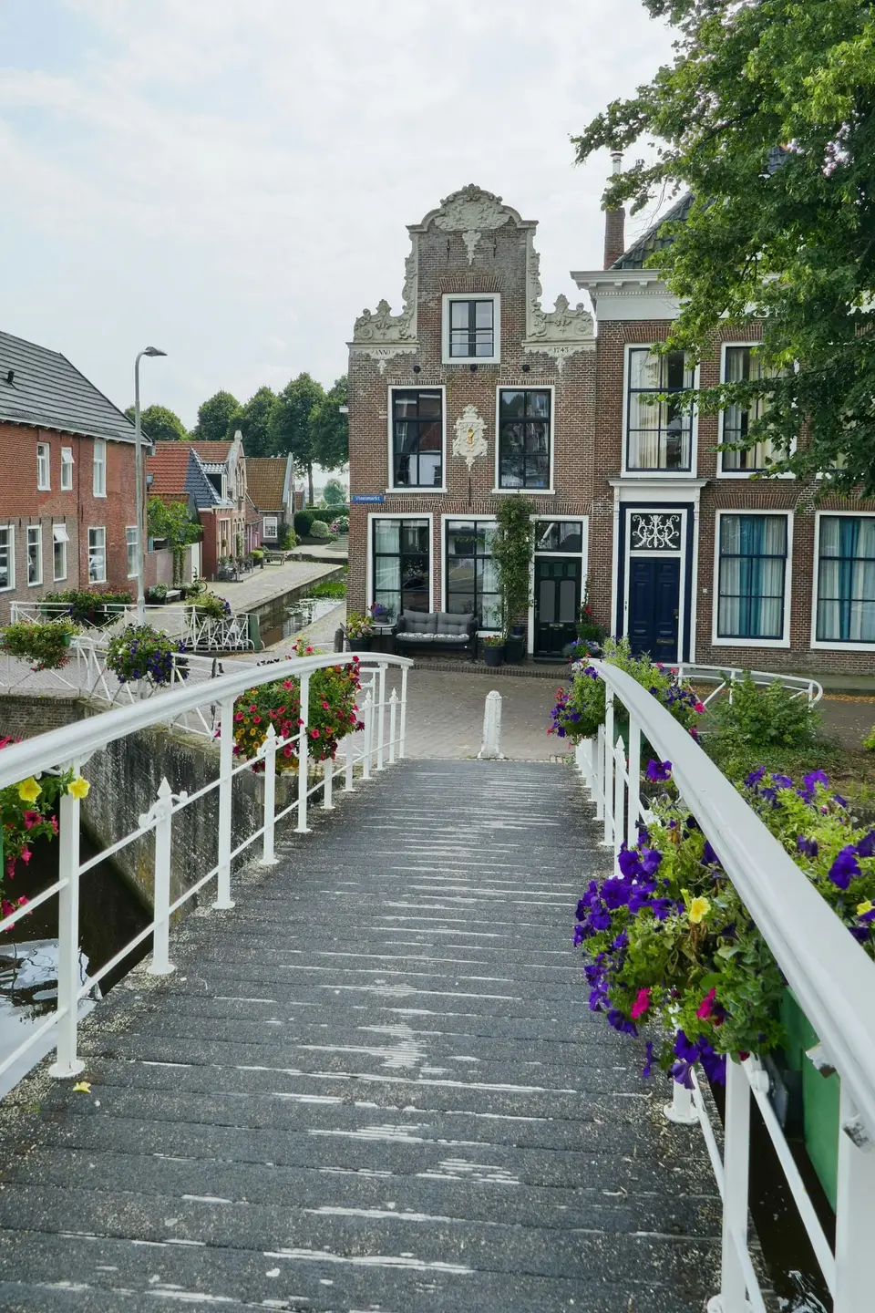 Witte voetgangersbrug met bloembakken in Dokkum, met uitzicht op een sierlijke gevel uit 1743 aan de Vleesmarkt